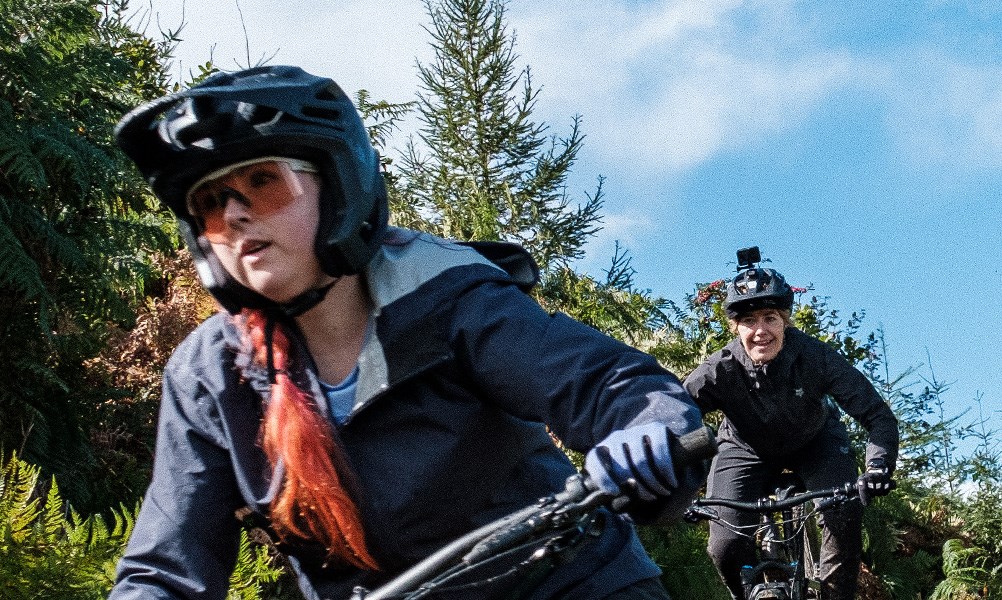 two female mountain bikers riding a trail wearing different MTB helmets